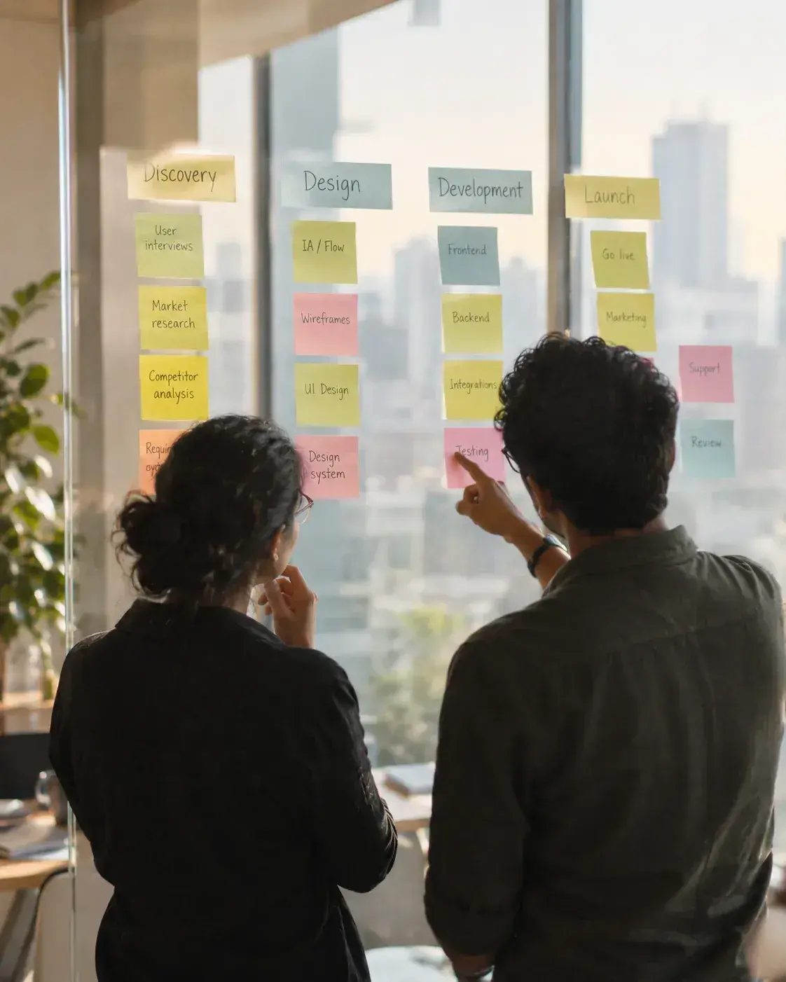 Two colleagues mapping stages of work across columns of sticky notes on a glass wall