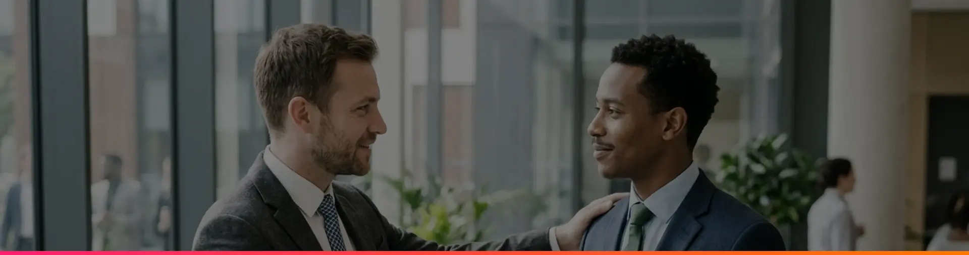 Two men in suits shake hands in a bright office lobby, one with a hand on the other's shoulder.