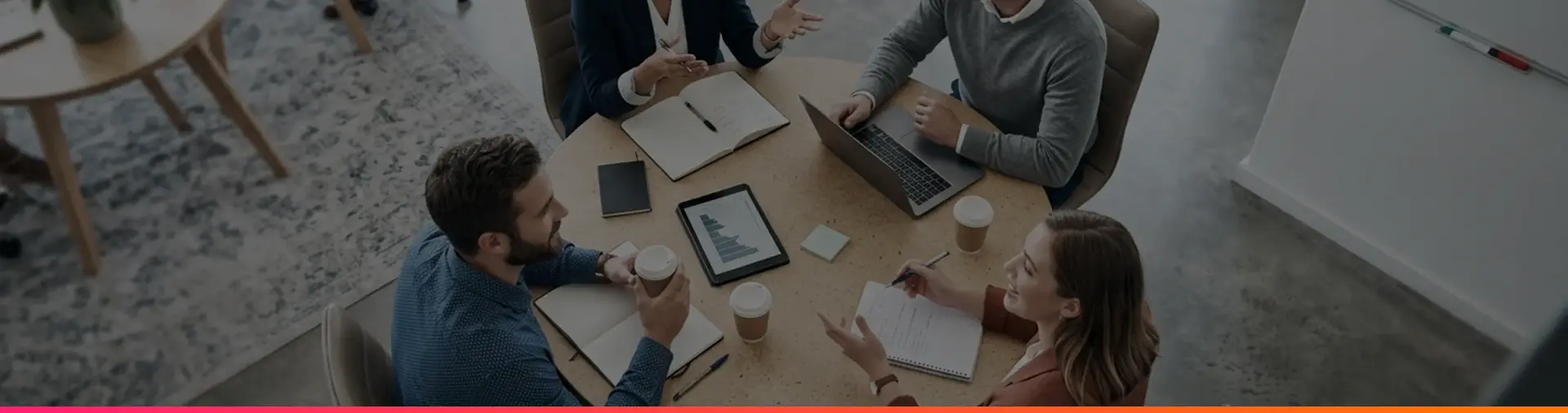 Four colleagues meet around a round wooden table with laptops, notebooks and coffee cups, seen from above